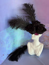 White ceramic bust sculpture used as a vase, holding three black roses and long black feathers against a textured pastel backdrop.