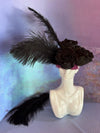 White ceramic bust sculpture used as a vase, holding three black roses and long black feathers against a textured pastel backdrop.