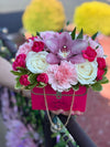 Hot pink purse box with chain handle holding cream roses, pink carnation, fuchsia spray roses, a purple orchid, and variegated greenery.