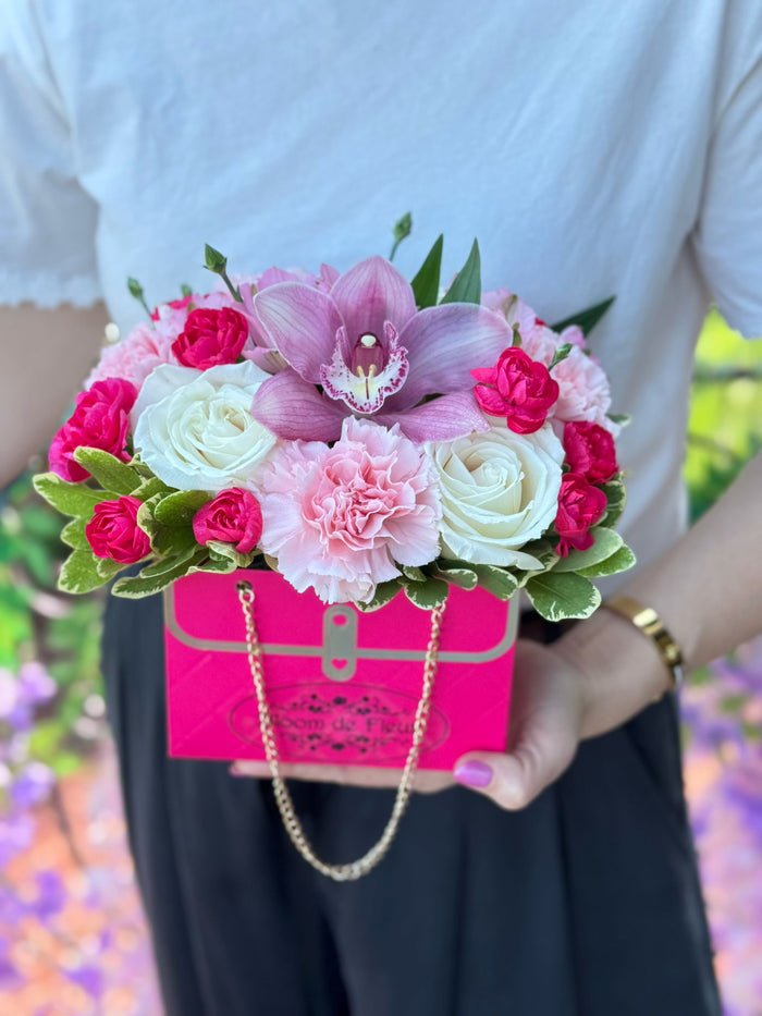 Hot pink purse box with chain handle holding cream roses, pink carnation, fuchsia spray roses, a purple orchid, and variegated greenery.