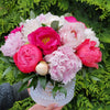 A pair of hands holding a white round “Bloom de Fleur” hatbox—adorned with a string of faux pearls and a gold scrollwork logo—overflowing with lush hot‑pink, coral, and pale blush peonies nestled in glossy green leaves, set against a backdrop of dense evergreen foliage.