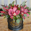 A matte black round gift box with a gold “Bloom de Fleur” logo and black rope handles sits on a wood‐grain tabletop, overflowing with large mauve‑pink cymbidium orchids, silvery eucalyptus leaves, and slender deep burgundy foliage; in the background, a black chair and a pale wall striped in cream and gray add subtle contrast.