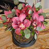 A matte black round gift box with a gold “Bloom de Fleur” logo and black rope handles sits on a wood‐grain tabletop, overflowing with large mauve‑pink cymbidium orchids, silvery eucalyptus leaves, and slender deep burgundy foliage; in the background, a black chair and a pale wall striped in cream and gray add subtle contrast.