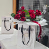 A white square paper gift bag with black ribbon handles and the word “FLOWER” printed in bold black letters holds a lush bouquet of deep red roses and silvery-green eucalyptus leaves, all wrapped in crisp white tissue paper and tied with a burgundy satin ribbon; in the softly blurred background, a second matching bag sits beside a window draped with beaded curtains and hanging silver snowflake ornaments.