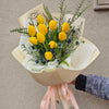 A wrapped bouquet of bright yellow tulips, spiky blue thistle blooms, delicate white filler flowers, and assorted greenery, all nestled in cream-and-light-green textured paper and tied with flowing pale pink ribbons against a neutral gray wall.