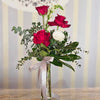 Clear tall glass cylinder vase tied with a soft gray satin ribbon, holding three deep red roses at varying heights, a single white ranunculus, tall white snapdragons, and sprigs of eucalyptus and mixed greenery, set on a wood tabletop against a pale striped background.