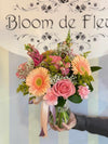 Clear glass vase tied with pink and lavender ribbons, holding peach gerbera daisies, pink roses, carnations, snapdragons, baby’s-breath, and solidago greenery.