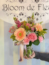 Clear glass vase tied with pink and lavender ribbons, holding peach gerbera daisies, pink roses, carnations, snapdragons, baby’s-breath, and solidago greenery.