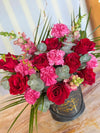 Black Bloom de Fleur hatbox with rope handles holding red roses, pink carnations, snapdragons, eucalyptus leaves, palm fronds.