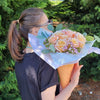 A person in a black knotted T‑shirt holds a waffle‑cone‑shaped Bloom de Fleur vessel wrapped in white and pale blue tissue paper, filled with peach‑cream roses, clusters of tiny lavender waxflowers, and eucalyptus sprigs, set against a backdrop of evergreen trees and pine‑straw ground.