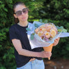 A person in a black knotted T‑shirt holds a waffle‑cone‑shaped Bloom de Fleur vessel wrapped in white and pale blue tissue paper, filled with peach‑cream roses, clusters of tiny lavender waxflowers, and eucalyptus sprigs, set against a backdrop of evergreen trees and pine‑straw ground.