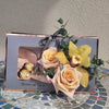 A rectangular gray Bloom de Fleur gift box with rose‑gold metallic trim sits on a mosaic tile table against a stucco wall; its left window reveals three gold‑foil wrapped chocolates, while the right side is decorated with two apricot roses, a yellow cymbidium orchid, eucalyptus sprigs, and a tied gray ribbon.